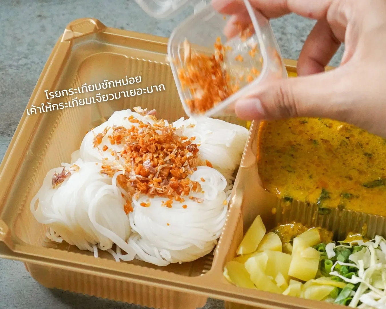โรยกระเทียมเจียวบนขนมจีน ข้างกล่องน้ำยาปูและผักเคียง