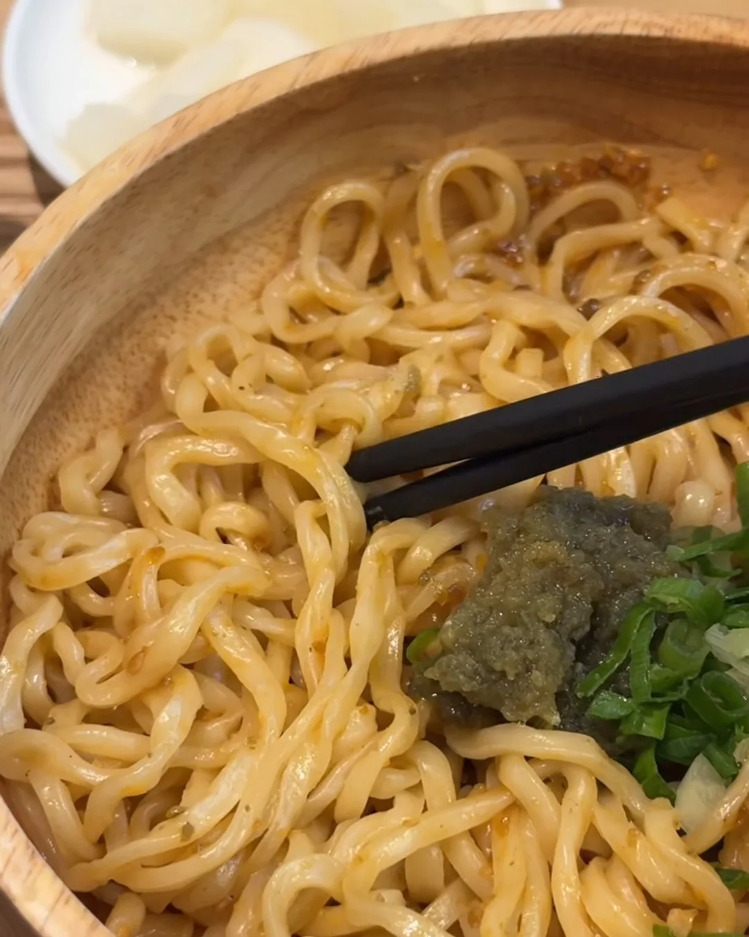 บะหมี่แห้งเส้นแบนหนึบในชามไม้ ตะเกียบคีบเส้น โรยต้นหอมและผักบดสีเขียว