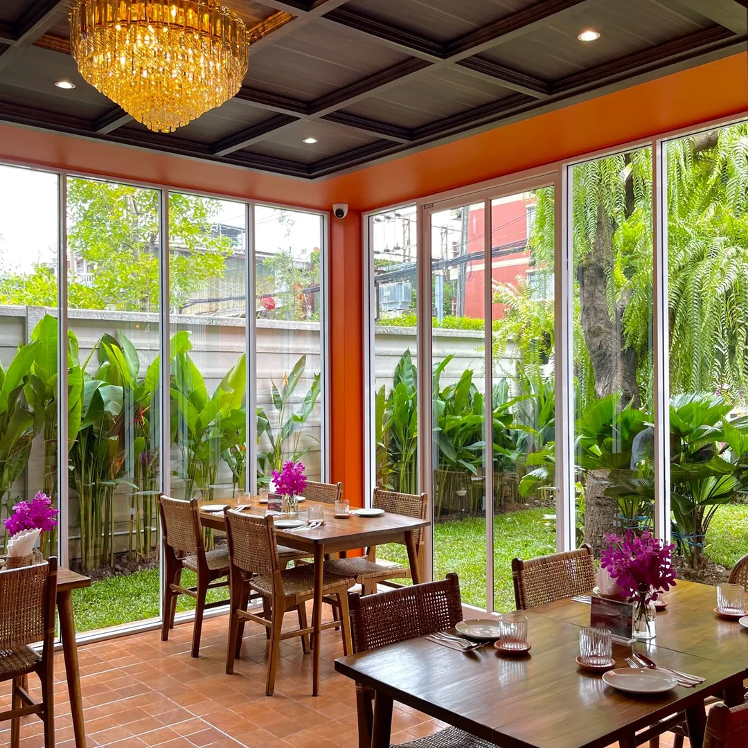 ห้องกระจกชั้นล่างมองเห็นสวนเขียว โต๊ะไม้และโคมคริสตัล ร้านปูนแดงแกงร้อน สาทร