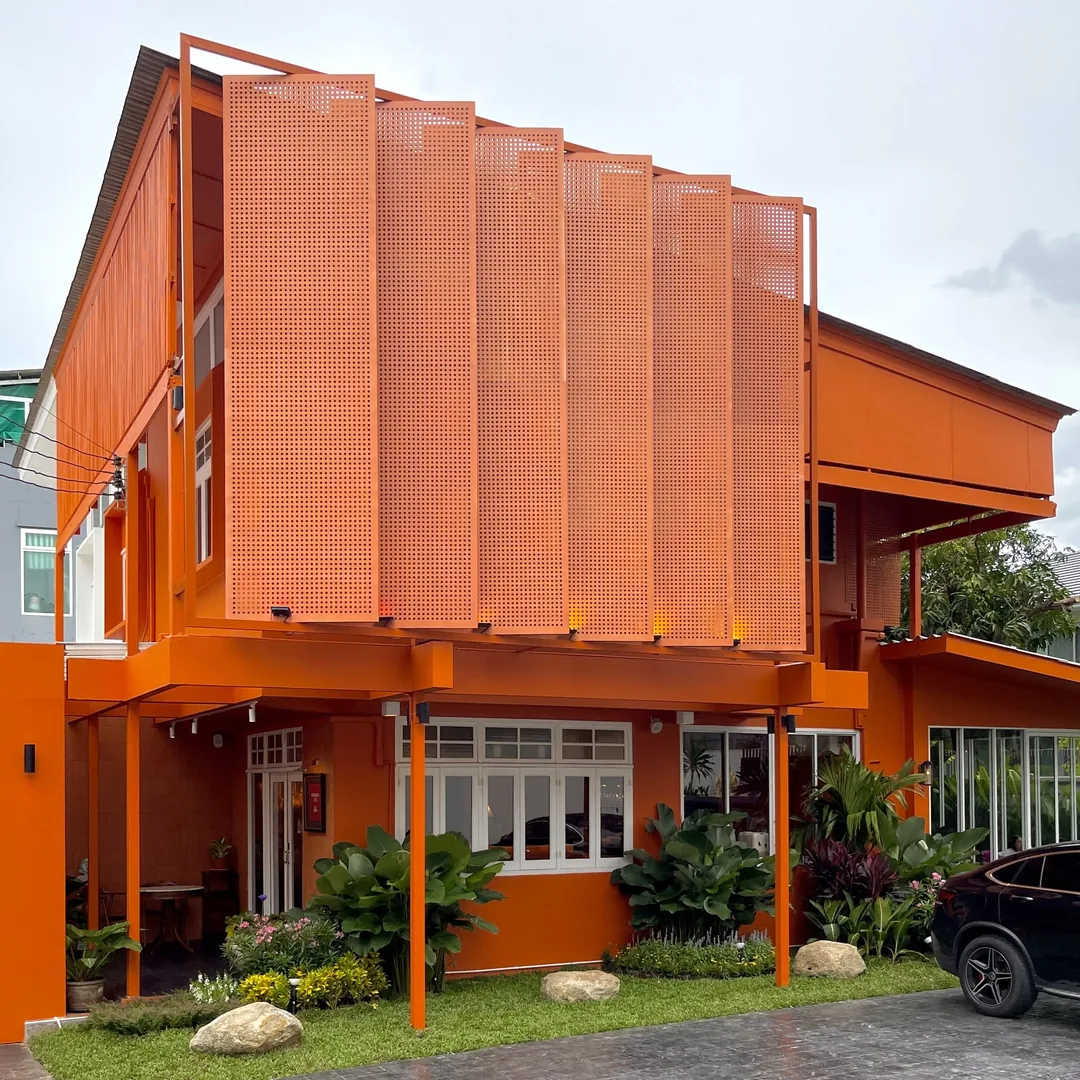 บ้านสองชั้นทาสีส้มอิฐ มีแผงระแนงเหล็กเจาะรูด้านหน้าและสวนเล็ก ๆ ร้านปูนแดงแกงร้อน สาทร