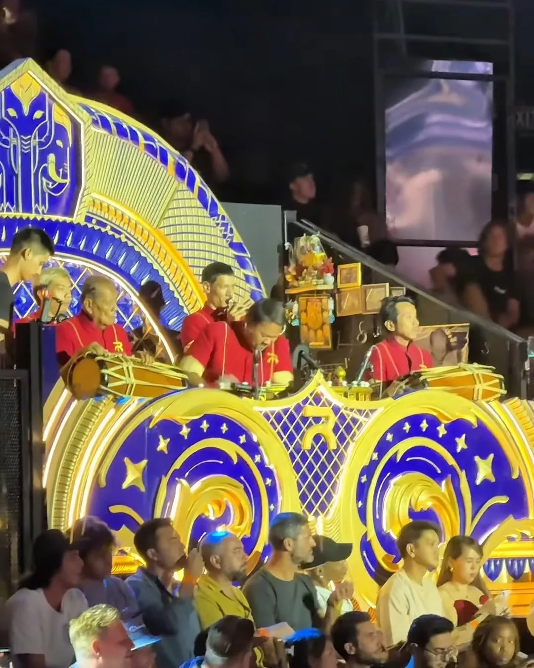 วงปี่กลองมวยไทยในเสื้อแดงบรรเลงสดบนซุ้มลายไทยสีน้ำเงินทอง เวทีมวยราชดำเนิน