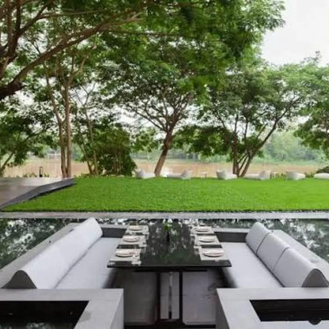 โซฟาสีเทาล้อมโต๊ะยาวกลางบ่อน้ำ มองเห็นสนามหญ้าและแม่น้ำกกที่ Riva Vista เชียงราย